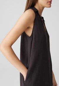 Mouwloze zwarte tuniek met een kraag, voorzien van een losse pasvorm en zijzakken. Zachte textuur en subtiele glans verbeteren het ontwerp.