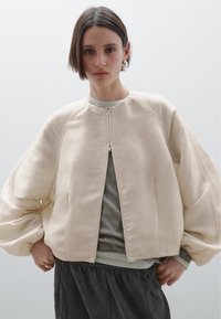 Jonge vrouw met kort haar, gekleed in een oversized crèmekleurige ritsjack over een grijze top en losse zwarte broek, tegen een effen achtergrond.