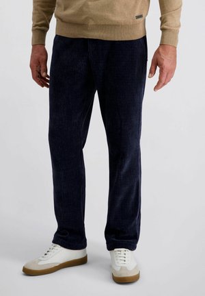 Man in een donkerblauwe corduroy broek, beige trui en witte sneakers met rubberen zolen, staande tegen een effen achtergrond.