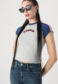 Camiseta gris de corte corto con mangas en azul y logotipo bordado en rojo "VERMIN". El modelo lleva gafas de sol angulares negras y jeans de talle alto en azul.