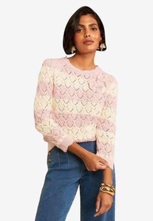 Frau trägt einen pink- und cremefarben gestreiften Strickpullover mit floralen Details und eine hoch taillierte blaue Jeans, ergänzt durch goldene Armbänder und große Ohrringe.