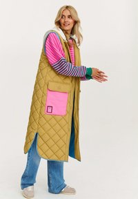 Prešívaná horčicová vesta s ružovou vreckom, položená na ružovom a zelenom pruhovanom tričku, v kombinácii s svetlo modrými džínsy a šedými teniskami.