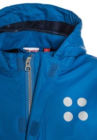 Giacca impermeabile blu con colletto alto, dettagli in punti argentati, chiusura con zip e toppe in Velcro sul davanti.