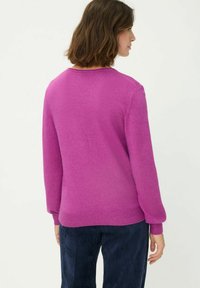 Leuchtend pinker Pullover aus weichem Material mit rundem Ausschnitt und geripptem Saum. Lange Ärmel, von hinten gezeigt, kombiniert mit marineblauen Cordhosen.