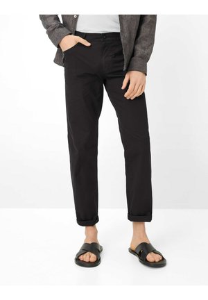 Uomo che indossa pantaloni neri risvoltati, una maglietta grigia con una mano in tasca e sandali neri a scivolo su sfondo bianco.