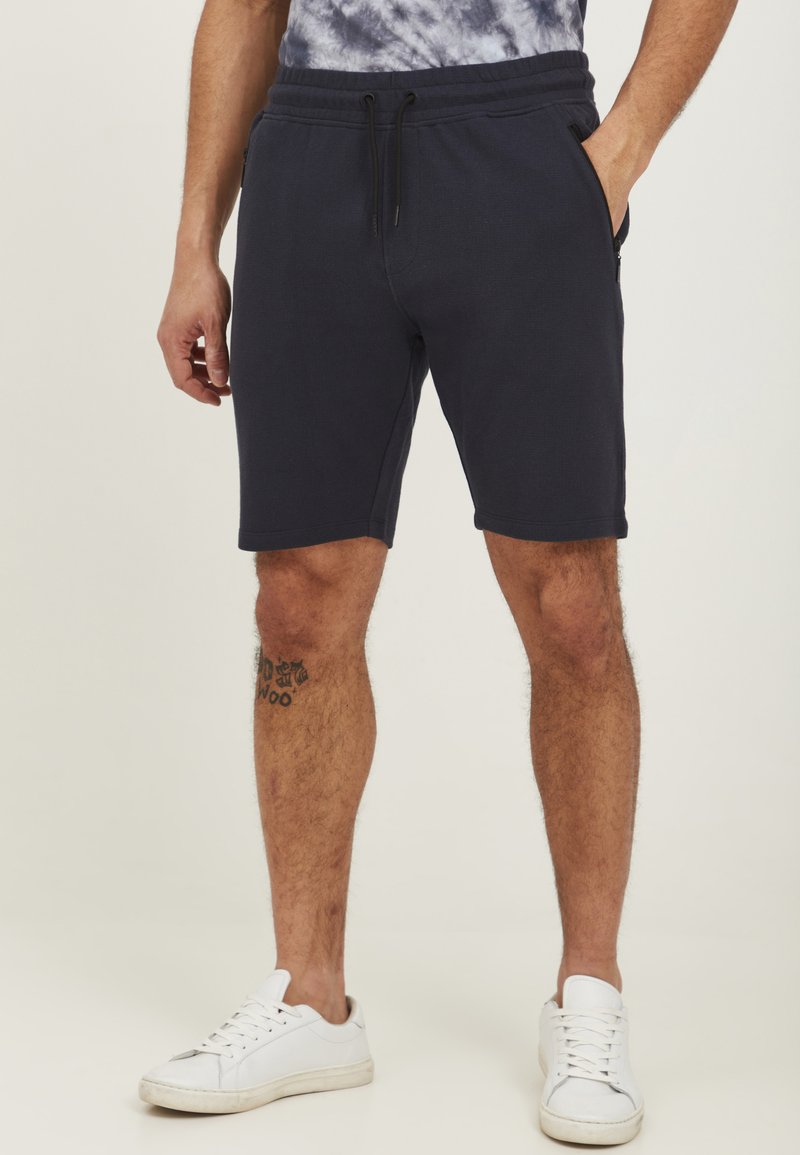 Donkerblauwe knielange shorts van een zachte stof, met een elastische tailleband en een trekkoord, en zijzakken. G gedragen met witte sneakers.