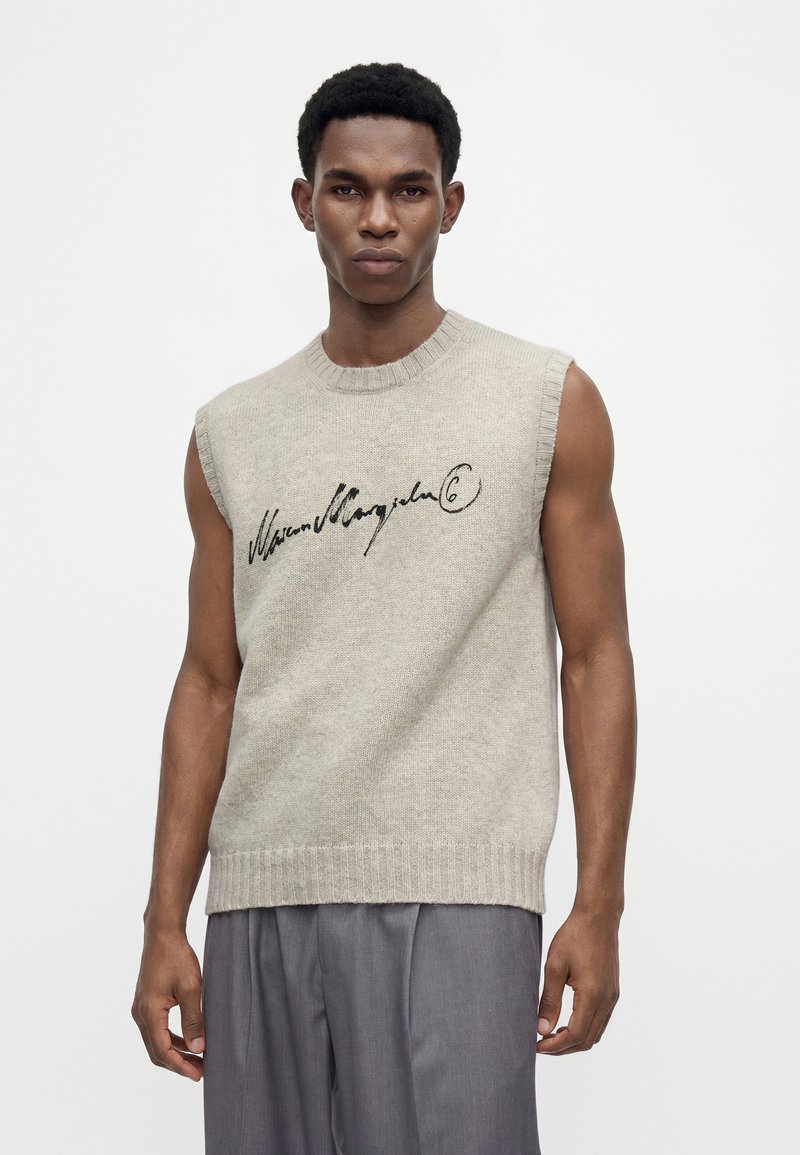 Mann mit kurzen Haaren, der einen ärmellosen beigen Strickpullover mit schwarzer Schrift über der Brust und lockere graue Hosen trägt, vor weißem Hintergrund stehend.