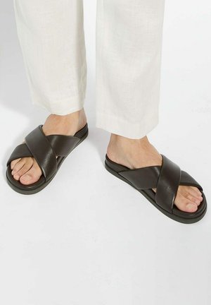 Brown leather slide sandals featuring a crossed strap design. Smooth texture and flat sole, suitable for casual wear.