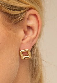 Boucles d'oreilles en métal doré avec un design rectangulaire creux. Texture lisse avec une finition brillante, créant une forme abstraite.