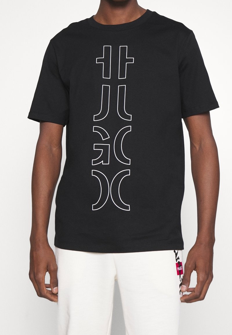 Homme portant un t-shirt noir à manches courtes avec un imprimé graphique vertical abstrait blanc et un short blanc avec un patch rose et noir.