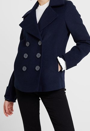 Navy blue double-breasted pea coat over white ribbed sweater, worn with black pants, hand in pocket, shown from neck to hips.