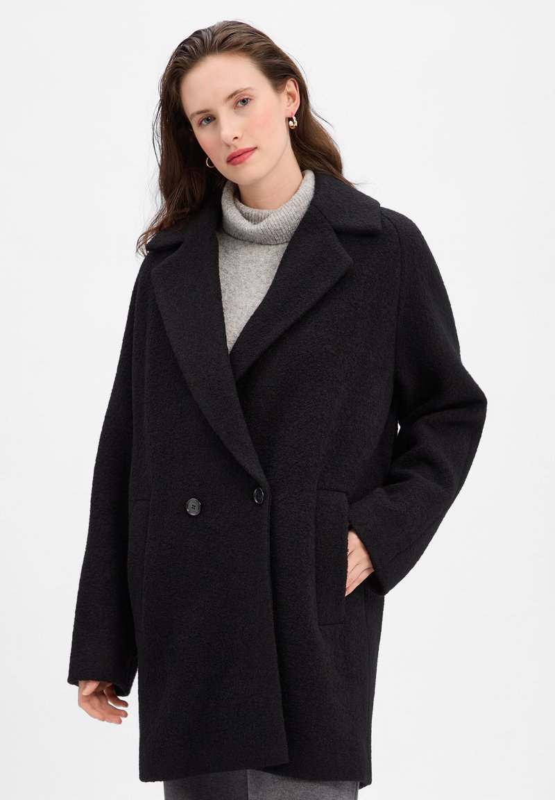 Manteau noir croisé en mélange de laine, avec de larges revers, des poches latérales et une coupe décontractée, porté sur un pull col roulé gris.