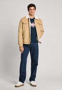 Traper jakna smeđe boje s bijelim krznom oko vrata, mornarskoplavi majica s motivom Union Jack, u kombinaciji s mornarskoplavim hlačama i bijelim tenisicama.