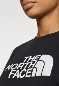 Μαύρο μπλουζάκι με μεγάλο λευκό λογότυπο που γράφει "THE NORTH FACE." Έχει στρογγυλή λαιμόκοψη και κοντά μανίκια με λεία υφή.