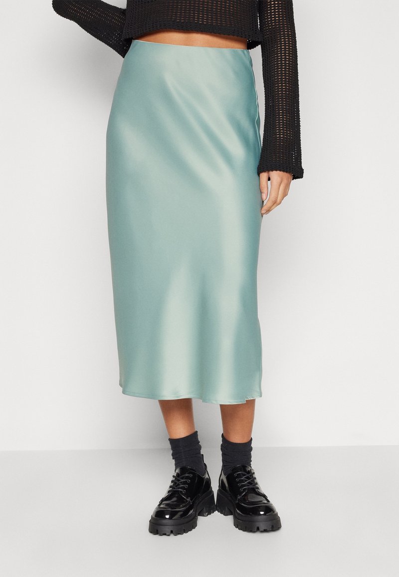 Femme portant une jupe midi en satin bleu clair, des chaussures en cuir noires épaisses avec des chaussettes noires, et un haut en mesh noir à manches longues.