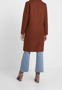 Brun uldfrakke, lige snit med lange ærmer. Parret med lyseblå flared jeans og teksturerede brune hælede støvler.