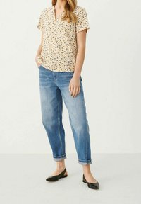 Blomstermønstret bluse med korte ærmer og v-udskæring i gul, parret med lyseblå, lige bukser; sorte flade sko fuldender outfittet.
