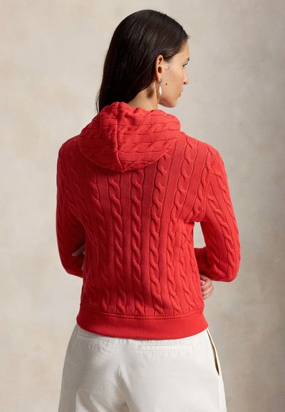 Vrouw met lang donker haar, gekleed in een rode kabelgebreide hoodie en witte broek, met gekruiste armen, met haar rug naar de camera toe.