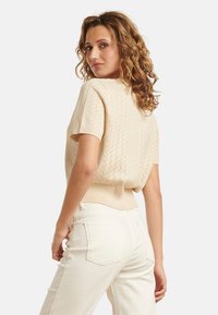 Korte mouw, beige gebreide top met een textuurpatroon en een geribbelde zoom. Gepaarde met lichtgekleurde, rechte broek.