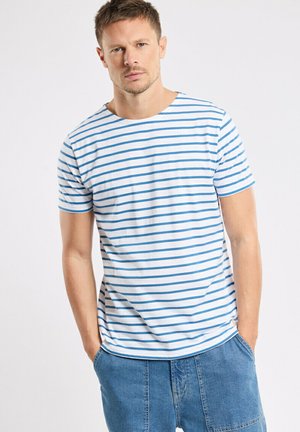 Homme portant un T-shirt blanc à manches courtes avec des rayures horizontales bleues et un pantalon en denim bleu, debout les mains dans les poches devant un fond uni.