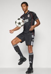 Juoda futbolo apranga su raudonomis detalėmis, susidedanti iš marškinėlių su apykakle ir šortų, derinama su juodais futbolo batais ir kelių aukščio juodomis kojinėmis su baltais dryžiais.