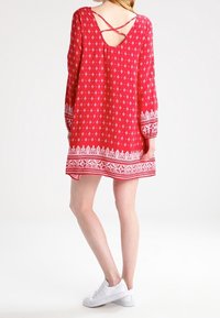 Robe rouge à motifs avec manches longues, dos en V et bordure blanche au bas. Associée à des baskets blanches.