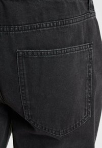 Los pantalones vaqueros de mezclilla negra presentan un bolsillo trasero con costuras tonal. La tela tiene una textura suave y un brillo sutil, destacando su diseño casual.