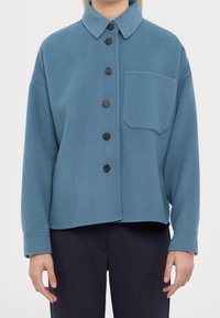 Giacca camicia in pile blu con colletto, sei bottoni scuri e taschino sul petto. Caratterizzata da una vestibilità rilassata e dettagli di cucitura a vista.