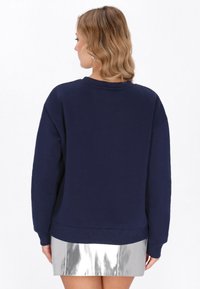 Tengerészkék pulóver kerek nyakkal és hosszú ujjal, laza szabással és bordázott szegéllyel. Ezüst metál miniszoknyával párosítva.