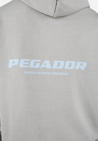 Lys grå hættetrøje med en blød tekstur. Har stor blå tekst "PEGADOR" på ryggen, med "CERTIFICERET AUTENTISK STREETWEAR" under.