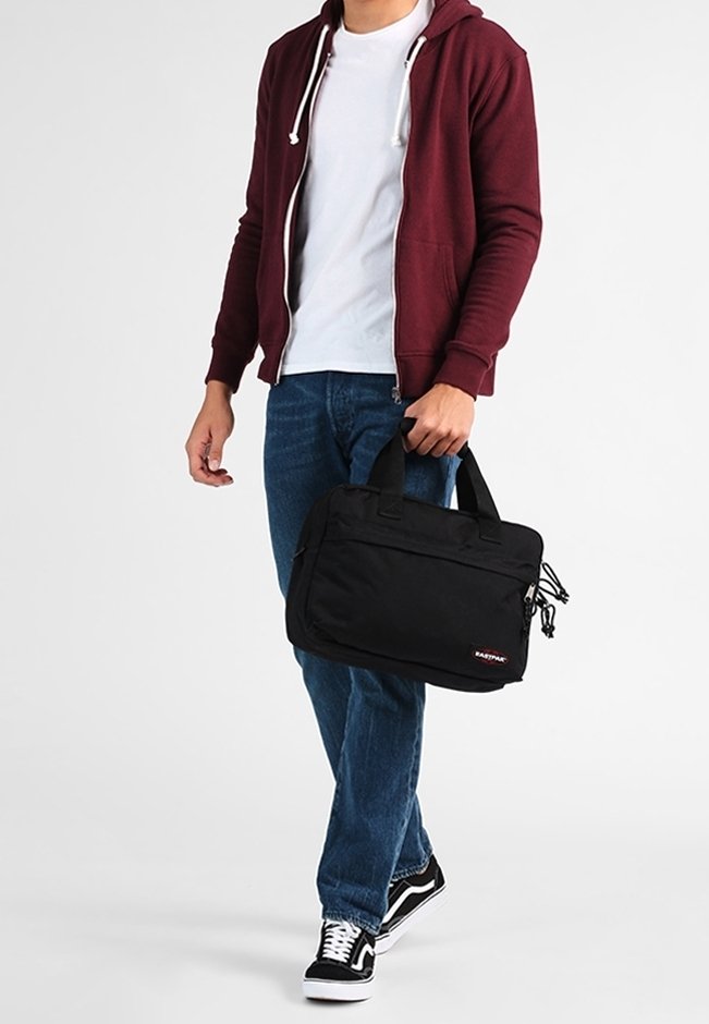 Personne portant un sweat à capuche bordeaux, un t-shirt blanc, un jean bleu et des baskets noires tenant un sac pour ordinateur portable Eastpak noir par les poignées.