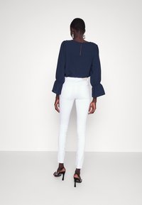 Blusa azul marino con mangas abullonadas y detalle de ojal en la espalda, combinada con jeans skinny blancos y sandalias de tacón alto negras con tiras.