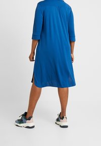 Woman standing with back facing, wearing a knee-length blue dress with three-quarter sleeves and colorful sneakers on a white background.