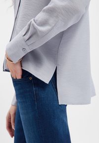 Light gray textured shirt with rolled sleeve, featuring a button closure and side slits, paired with dark blue denim jeans.