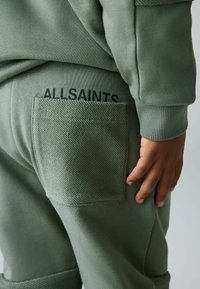 Joggers de algodón verde con tejido texturizado, que presentan un bolsillo parche en la parte posterior y un logo impreso, mostrados desde el lado y la parte trasera.