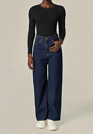 Person wearing a black long-sleeve top, high-waisted wide-leg dark blue jeans, and white sneakers, standing with one hand on the waist.