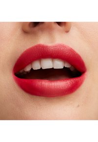 Jasnoczerwona matowa szminka na pełnych ustach, pokazująca gładką teksturę i wyraźne kontury, z białymi zębami widocznymi w tle.