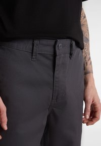Un pantalon en coton gris foncé doté d'une fermeture à bouton, de passants de ceinture et de poches latérales. Texture lisse avec des détails de couture minimaux.