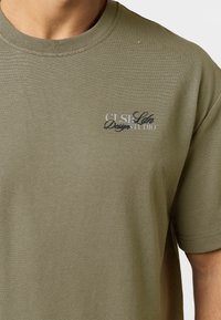 Άνδρας που φορά ελιάς πράσινο κοντομάνικο T-shirt με μικρό, επικαλυπτόμενο ασπρόμαυρο λογότυπο κειμένου στο αριστερό στήθος που γράφει "CLSP Ldn Design Studio."