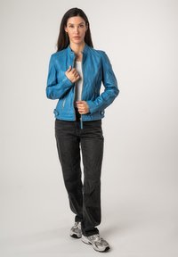 Hellblaues Lederjacke mit Stehkragen, vorderem Reißverschluss und seitlichen Taschen, kombiniert mit schwarzen Jeans und grauen Turnschuhen.