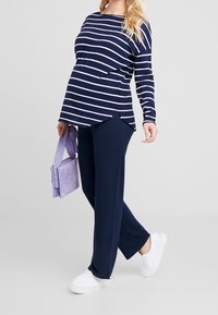 Femme enceinte portant un haut rayé bleu marine, un pantalon bleu marine, des baskets blanches et un sac à main lavande sur un fond clair.
