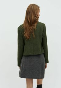 Veste verte en chevrons avec des épaules structurées, une coupe ajustée et des manches longues, associée à une jupe grise à motif pied-de-poule.