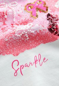 Biała tkanina z różowym gradientem, z akcentami cekinów i koralików w kształcie motyli oraz napisem "Sparkle" w różowym kursywie.