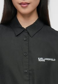 Svart skjorte med knappestolpe, spiss krage, teksturert stoff og brodert merke "KARL LAGERFELD JEANS" i hvitt.