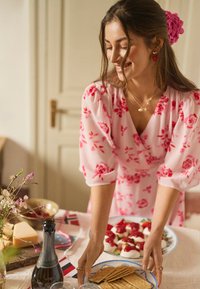 Žena v ružových kvetovaných šatách kladie tanier sušienok na ozdobený stôl so syrom, jahodovými jednohubkami a fľašou šumivého vína.