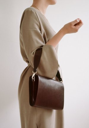 Kvinde i beige kjole holder brun læderhåndtaske i den korte rem med hånden hævet nær brystet mod en ensfarvet lys baggrund.