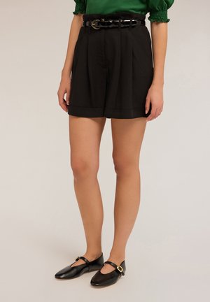 Sorte plisserede shorts med en bred talje, der har et sort flettet bælte. Tekstureret stof, opsmøg ved kanten, parret med sorte lædersko.