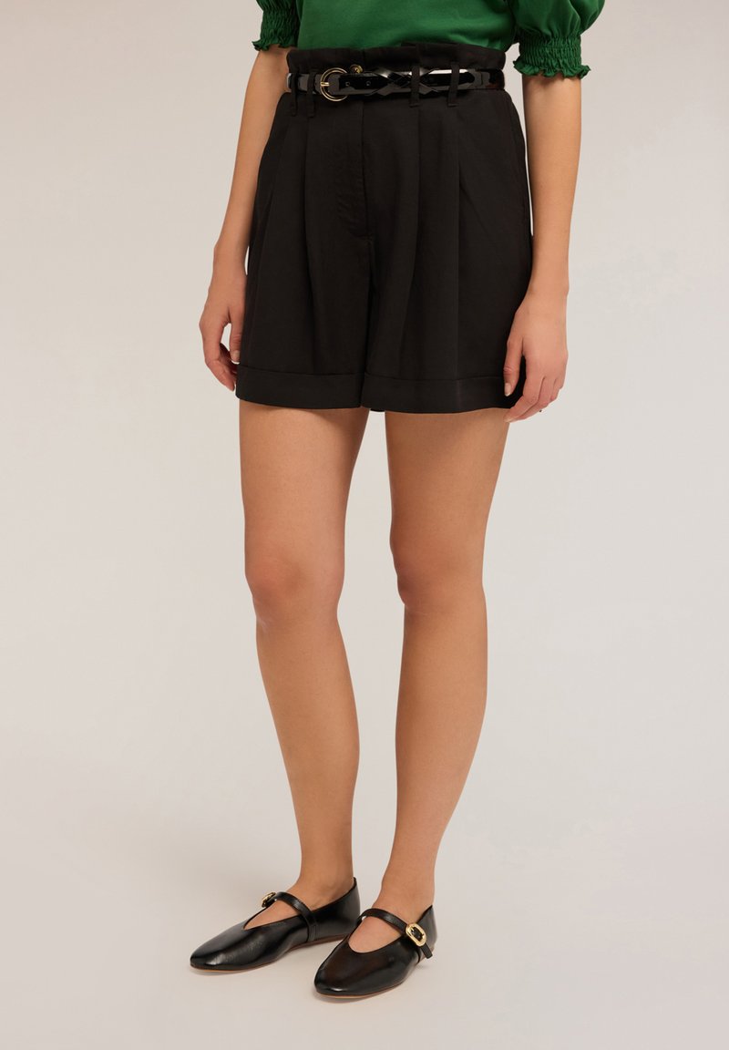 Mustat pleatatut shortsit leveällä vyötärönauhalla, jossa on musta punottu vyö. Teksturoitu kangas, käännetty helma, yhdistetty mustiin nahkakenkiin.