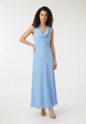 Donna con capelli ricci che indossa un vestito azzurro chiaro senza maniche, lungo fino al pavimento, e tacchi dorati aperti sulle dita, in piedi con una mano sul fianco.