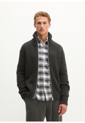 Uomo che indossa una giacca scura con zip e trama sopra una camicia a quadri bianca e nera con le mani nelle tasche.